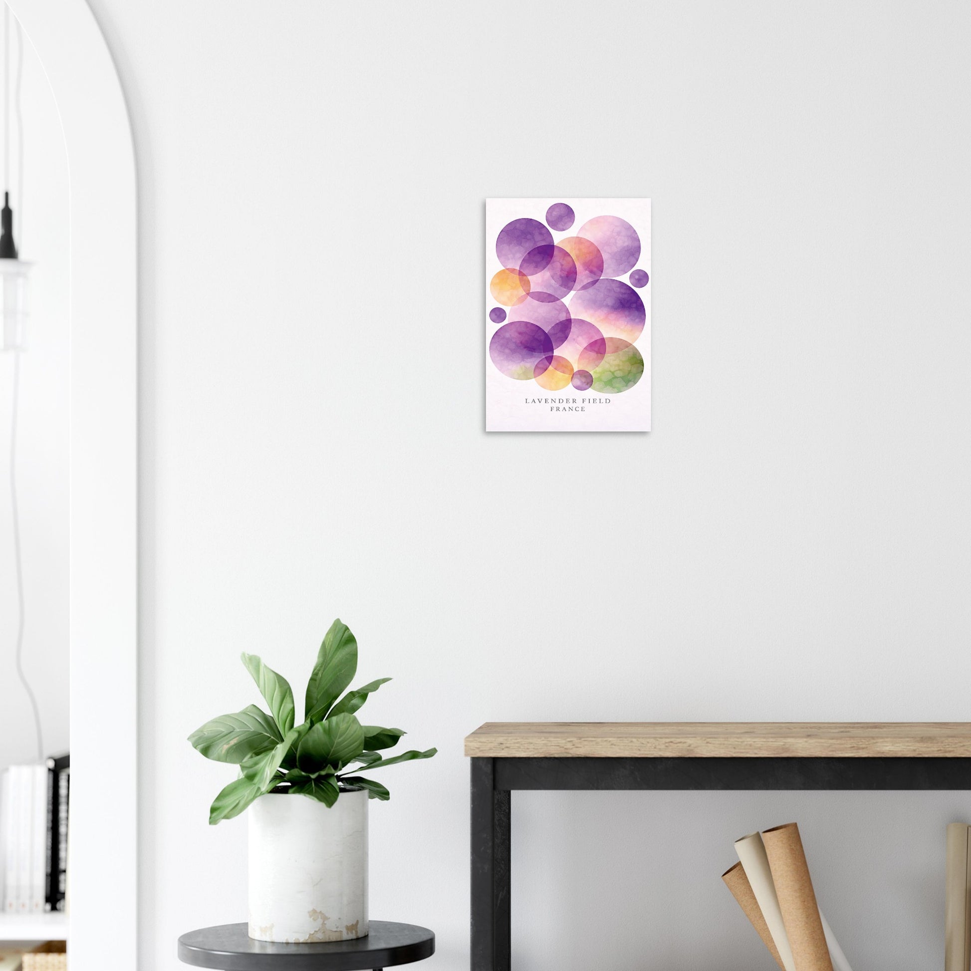 A framed wall art print featuring overlapping watercolor circles in purple, pink, orange, and green on a white background, displayed on a light gray wall above a wooden table with a potted plant nearby.