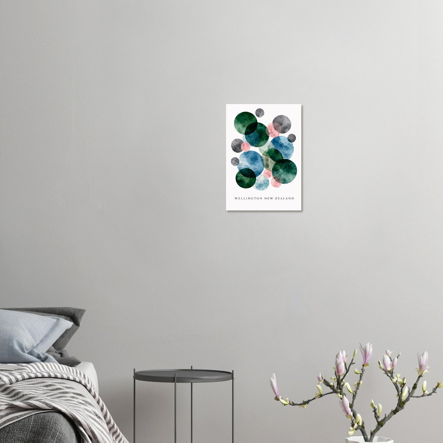 A framed wall art print featuring abstract green, blue, and gray watercolor circles on a white background, hung on a light gray wall above a bed with a small side table and flowering branches in the foreground.
