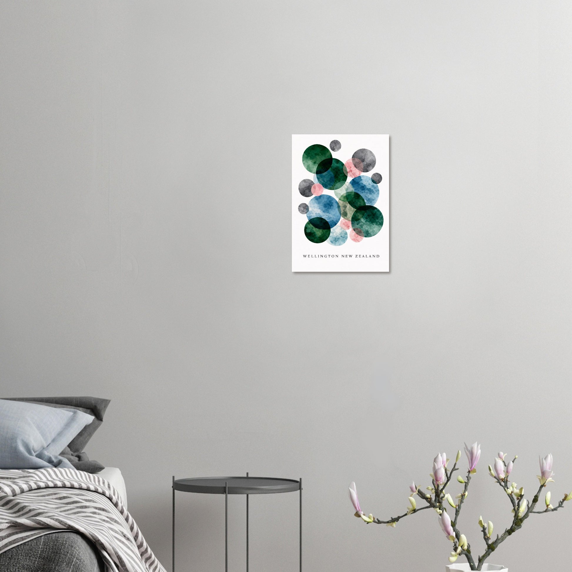 A framed wall art print featuring abstract green, blue, and gray watercolor circles on a white background, hung on a light gray wall above a bed with a small side table and flowering branches in the foreground.