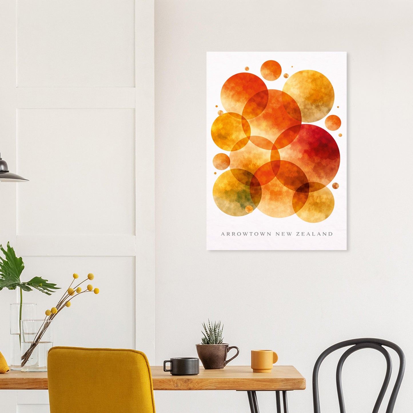 A vertical art print featuring overlapping watercolor circles in orange, yellow, and red tones, with the text 'ARROWTOWN NEW ZEALAND' at the bottom, displayed on a white wall above a dining table in a modern room setting.