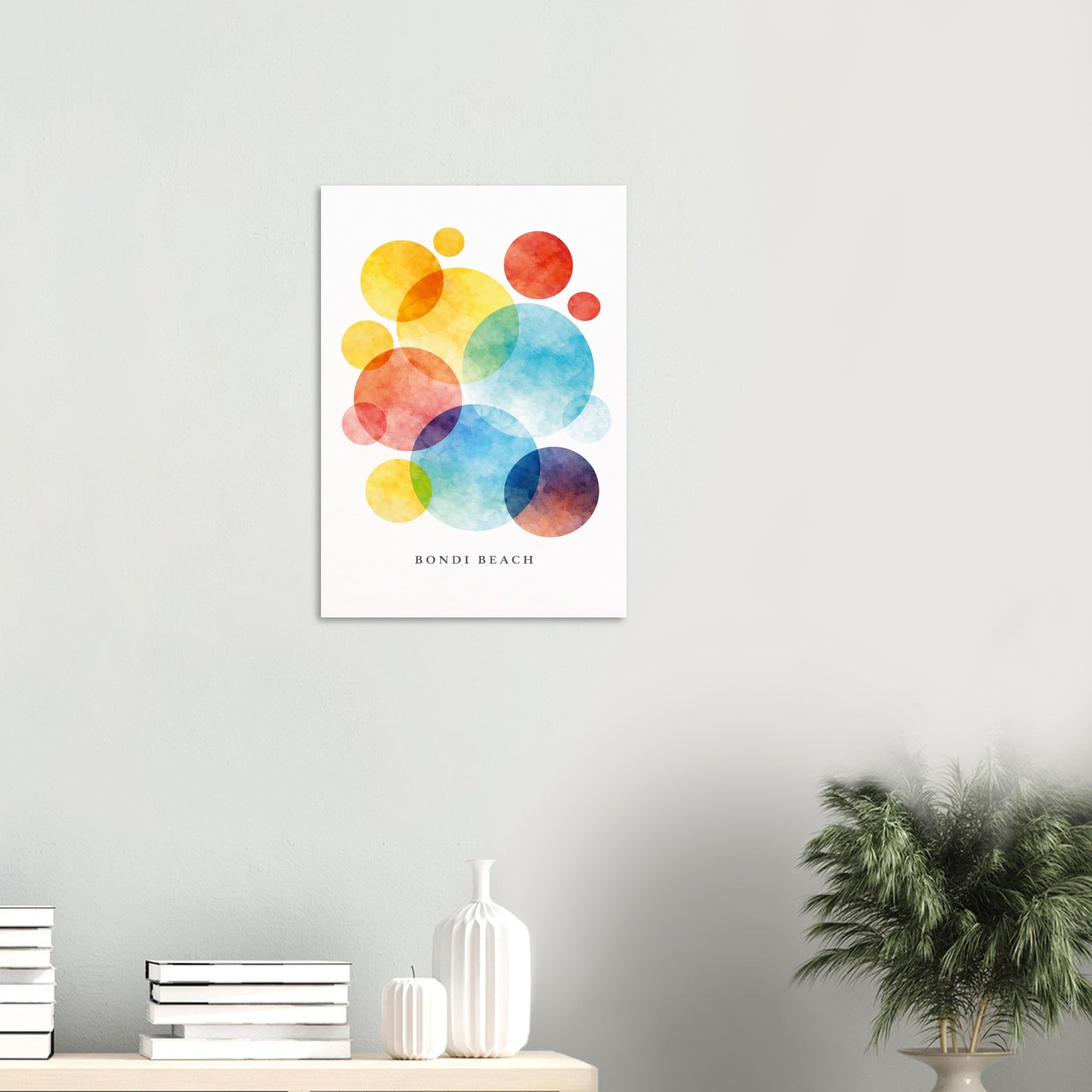 A vertical wall art print featuring colorful overlapping watercolor circles in yellow, orange, red, blue, and purple on a white background, with the text 'BONDI BEACH' printed below. The print is displayed on a light gray wall above a shelf with books, white vases, and a potted plant.