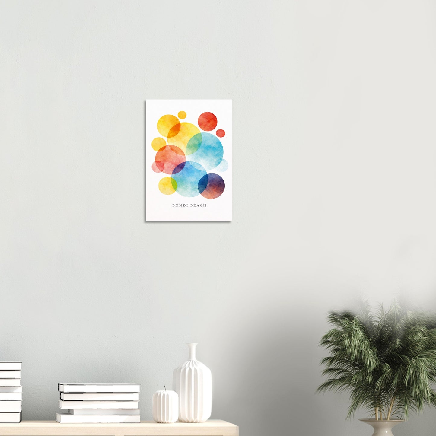 A vertical watercolor art print with colorful overlapping circles in yellow, orange, red, blue, and purple on a white background, hanging on a light gray wall above a shelf with books, vases, and a potted plant.