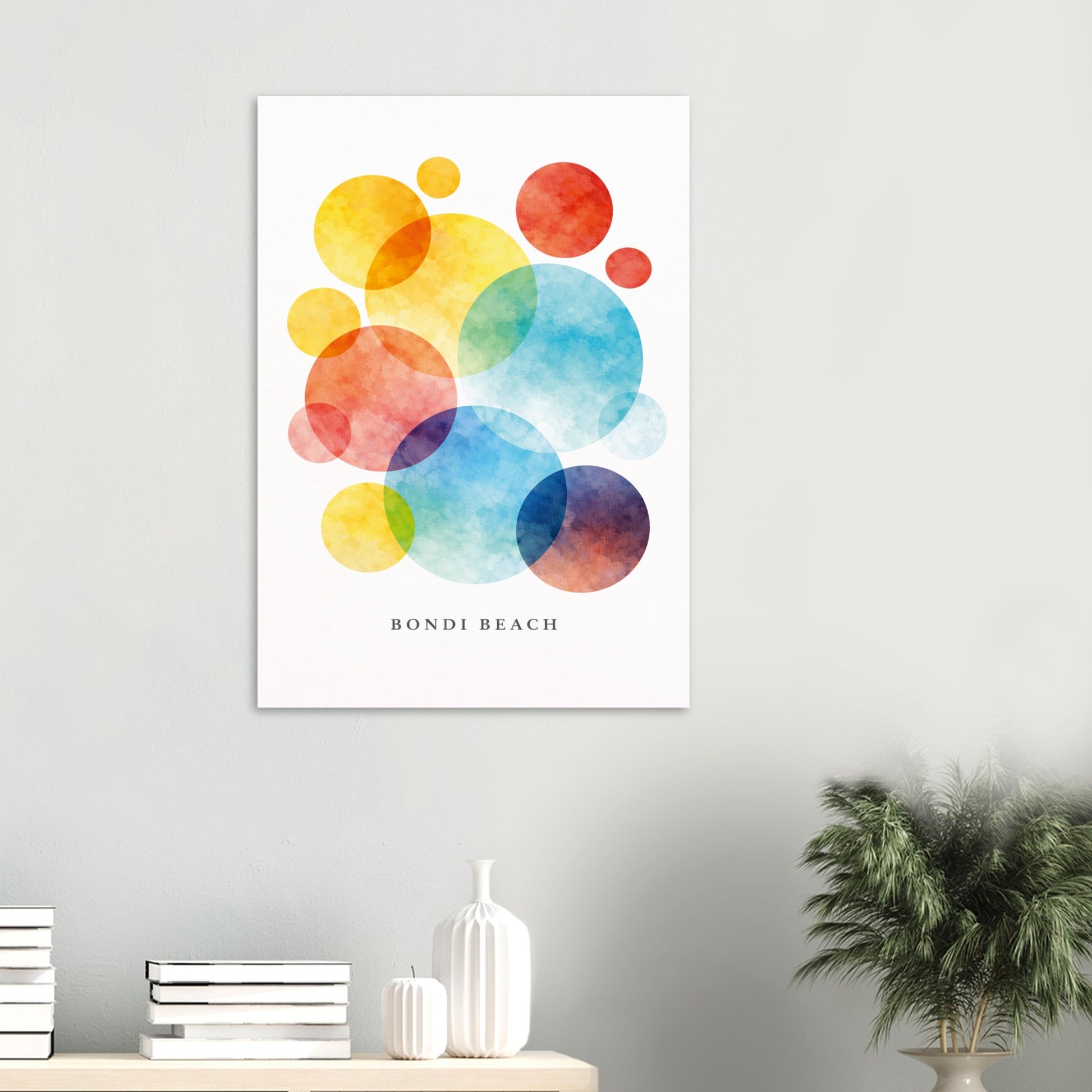 A vertical wall art print featuring colorful overlapping watercolor circles in yellow, orange, red, blue, and purple on a white background, with the text 'BONDI BEACH' near the bottom. The print is displayed on a light gray wall above a small table with books, a white vase, and a potted plant.