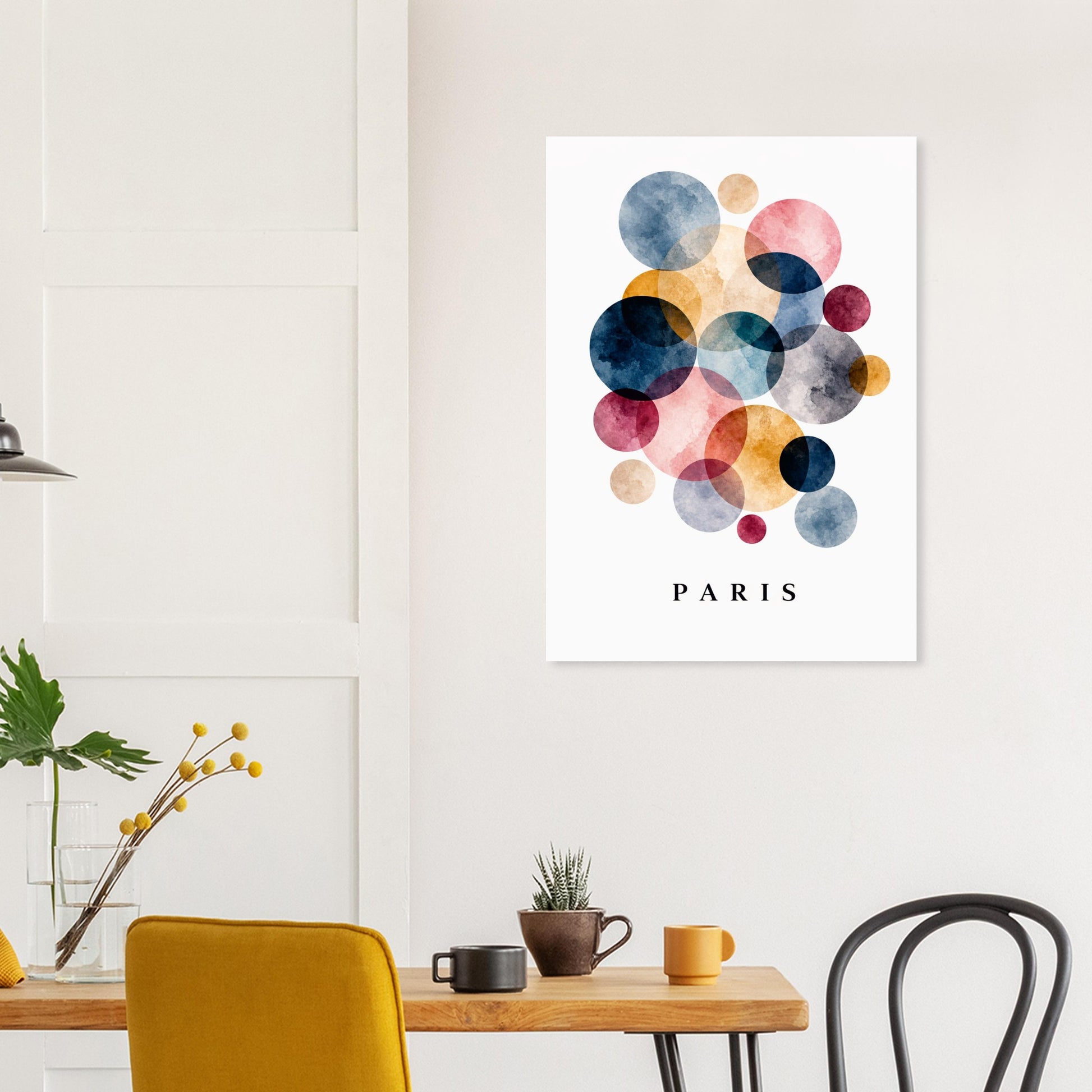 A vertical wall art print featuring overlapping watercolor circles in blue, pink, yellow, and orange on a white background, with the word PARIS printed below in black. The print is displayed in a modern dining room setting above a wooden table with chairs and small decor items.