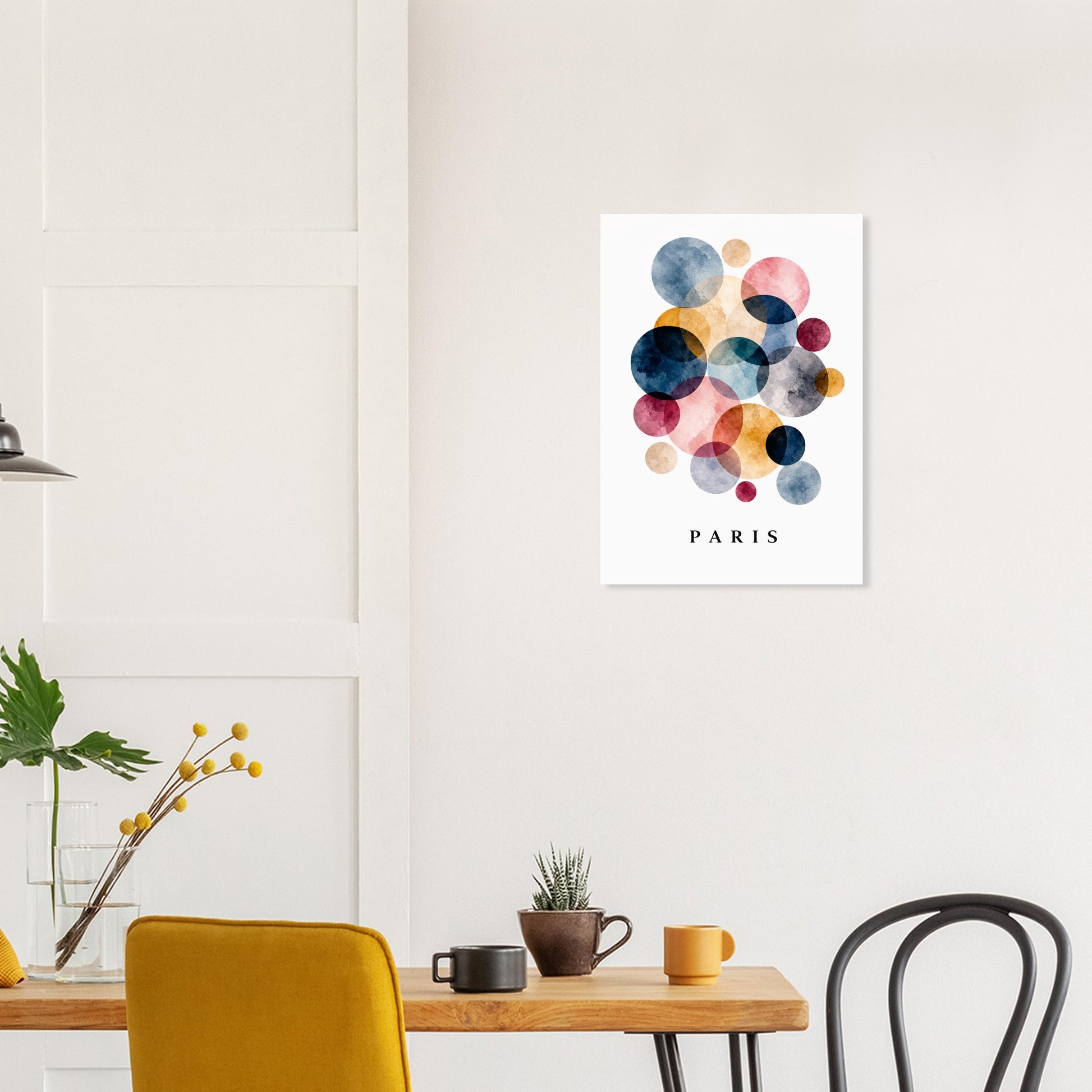 A vertical wall art print featuring colorful watercolor circles above the word PARIS, displayed on a white wall in a modern dining room setting with a wooden table, chairs, and plants.