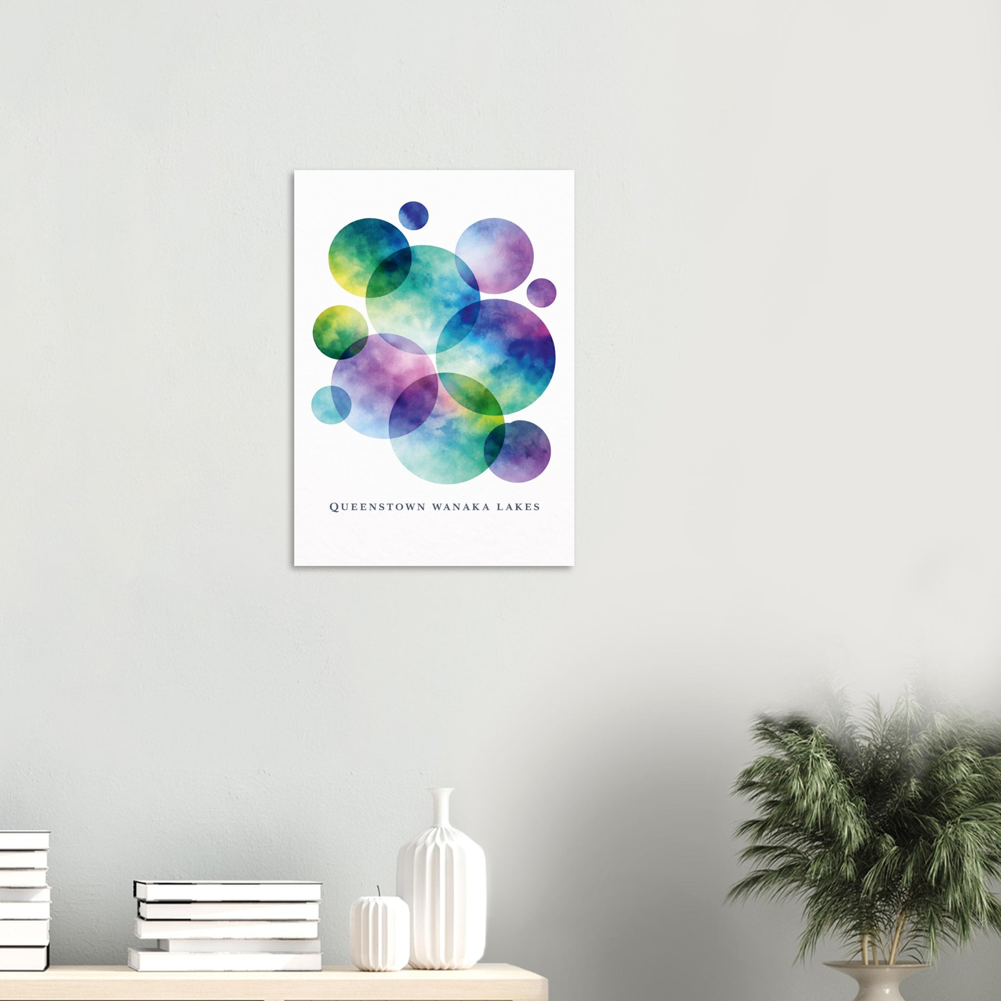 A vertical wall art print featuring colorful watercolor bubble shapes in blue, green, purple, and yellow on a white background, with the text 'QUEENSTOWN WANAKA LAKE' below. The print is displayed on a light gray wall above a shelf with books, a white vase, and a potted plant.