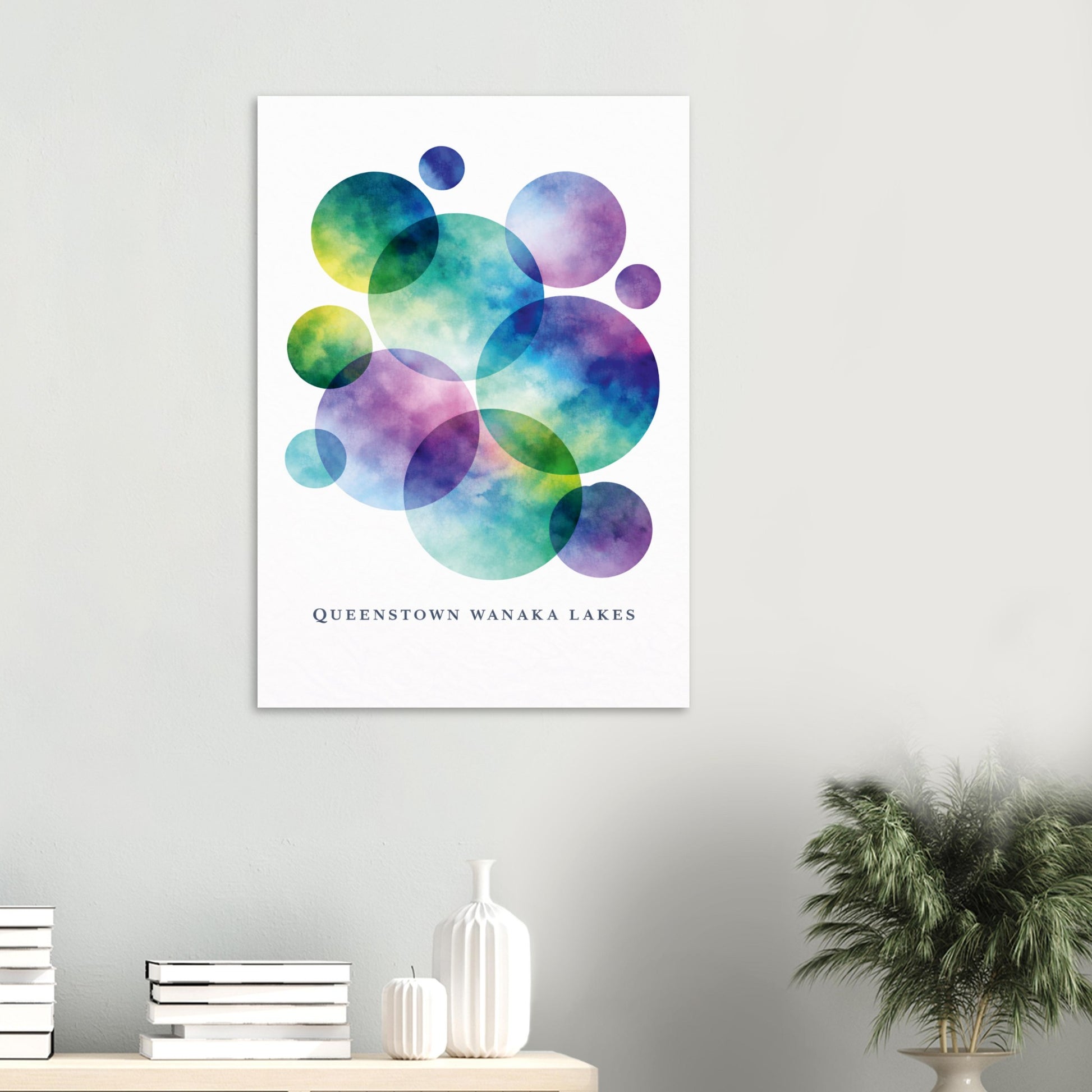 A vertical wall art print featuring overlapping watercolor circles in teal, blue, purple, and green on a white background, with the text 'QUEENSTOWN WANAKA LAKES' near the bottom. The print is displayed on a light gray wall above a console table with books, a white vase, and a potted plant.