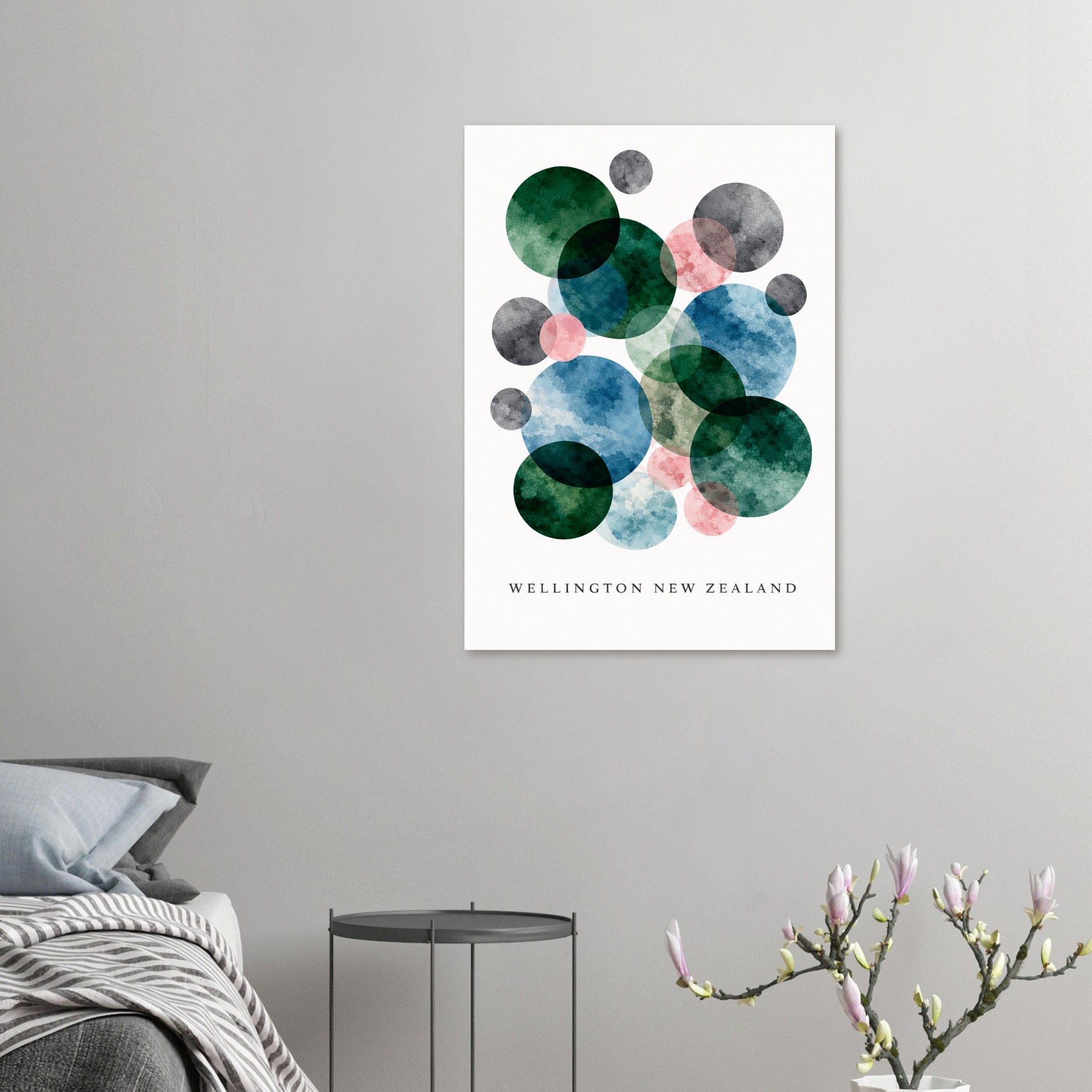 A vertical wall art print featuring overlapping watercolor circles in green, blue, pink, and gray on a white background, with the text 'WELLINGTON NEW ZEALAND' at the bottom. The print is displayed in a bedroom setting on a light gray wall above a bed and side table.