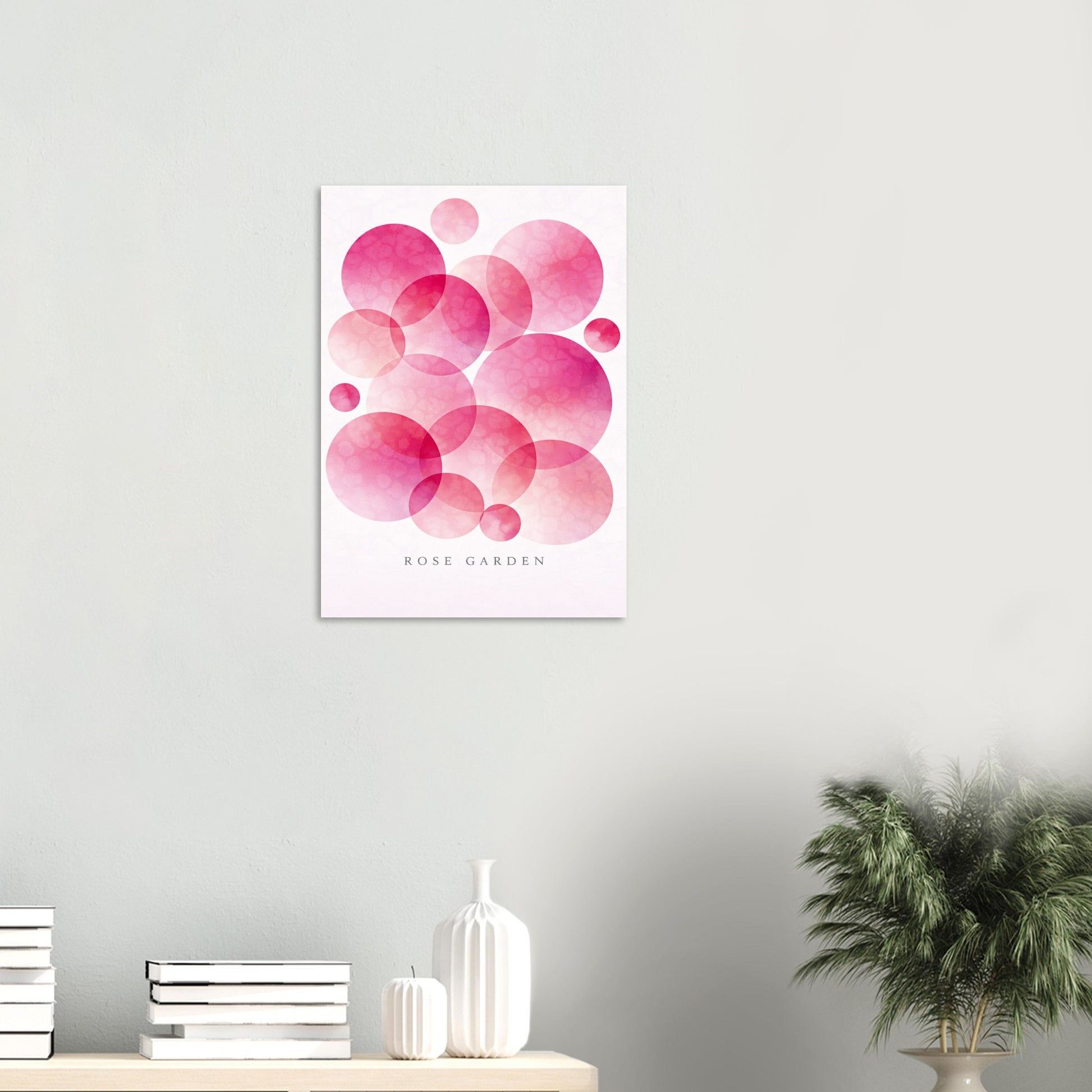 A vertical wall art print featuring overlapping pink and magenta watercolor circles on a white background, with the text 'ROSE GARDEN' near the bottom. The print is displayed on a light gray wall above a shelf with books, white vases, and a potted plant.