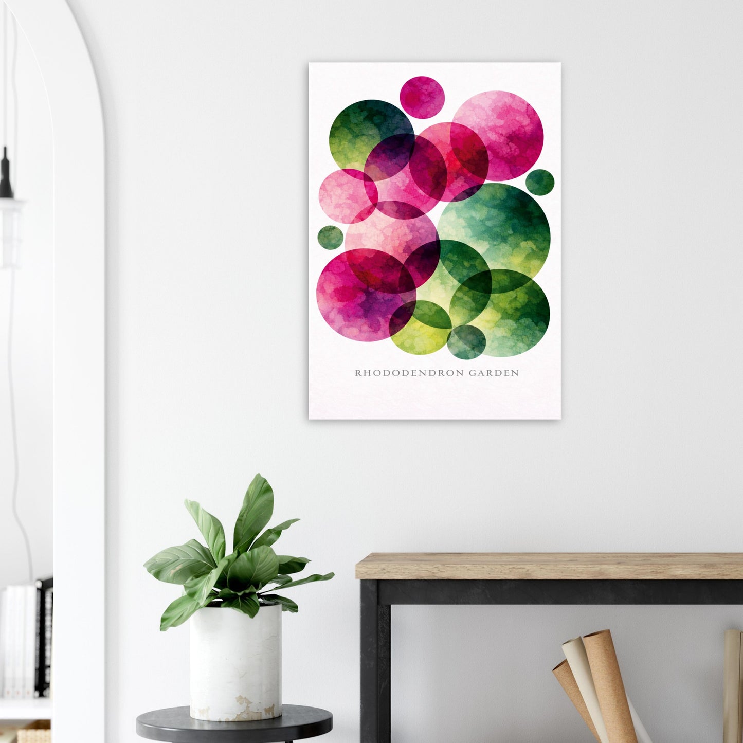 A vertical wall art print featuring overlapping translucent watercolor circles in pink, green, and yellow on a white background, with the text 'RHODODENDRON GARDEN' near the bottom. The print is displayed in a styled interior setting above a wooden table and next to a potted plant.
