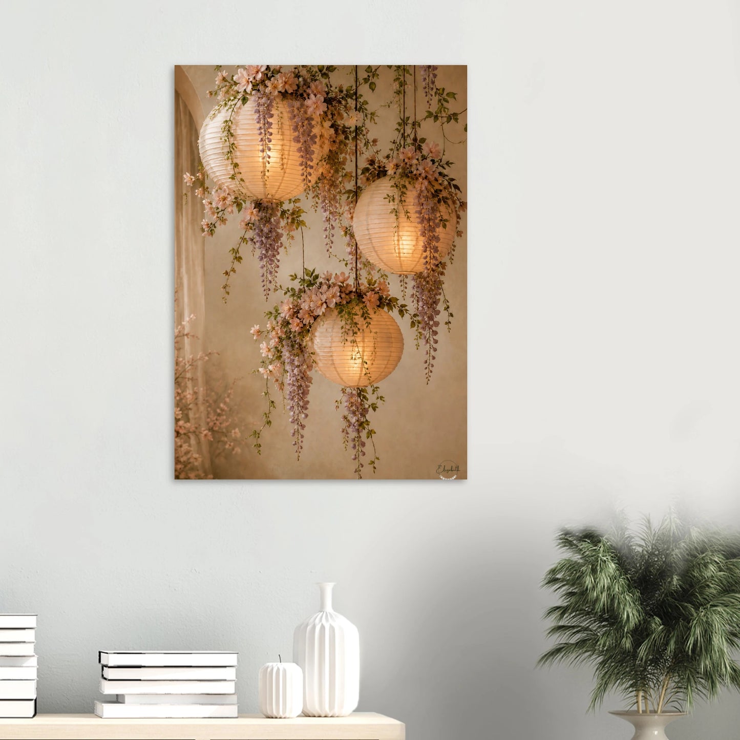 A vertical wall art print depicting three glowing paper lanterns and wisteria flowers, displayed on a light gray wall above a console table with books, a vase, and a potted plant.