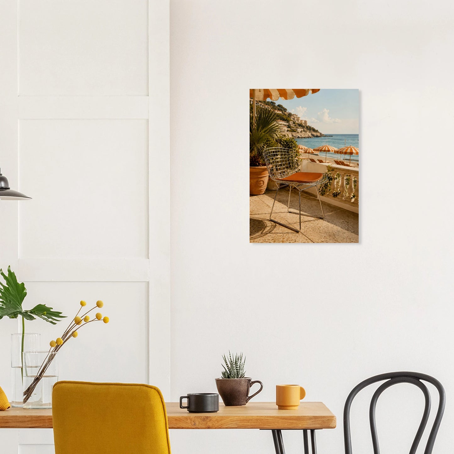 A vertical canvas wall art print depicting a sunny coastal patio scene with palm trees, an orange chair, and the blue sea, displayed on a white wall above a dining table in a modern room setting.