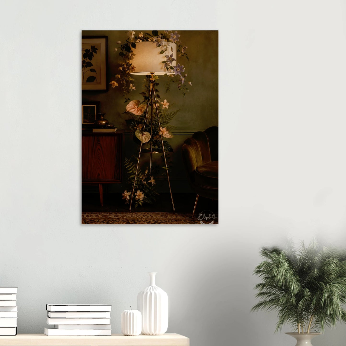 A vertical wall art print depicting a moody, vintage-style interior with a tripod floor lamp, floral garlands, and a wooden cabinet, displayed on a light gray wall above a console table with decorative vases and a potted plant.