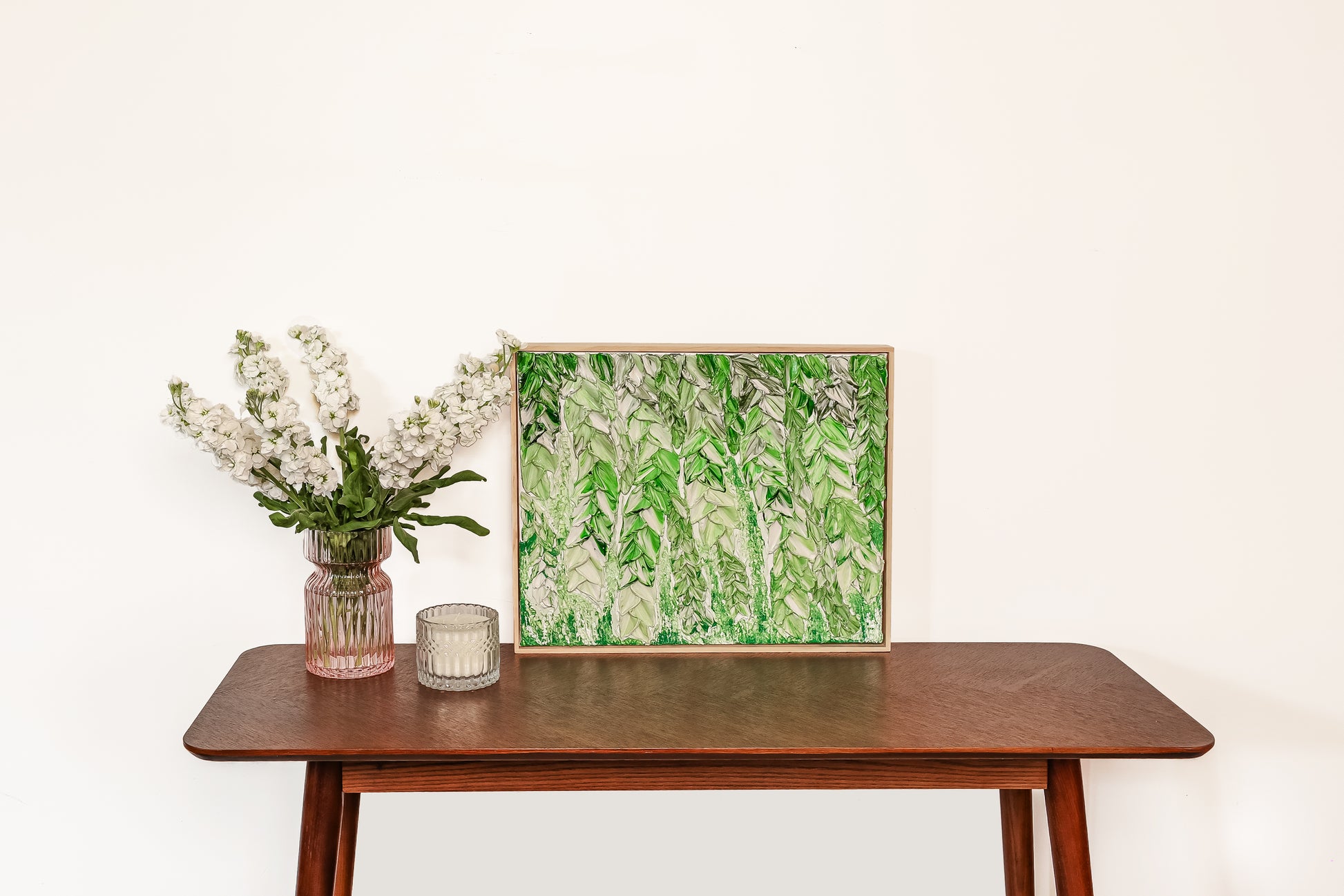 A framed botanical art print featuring green and white leafy patterns, displayed on a wooden console table next to a pink glass vase with white flowers and a small candle holder against a plain white wall.