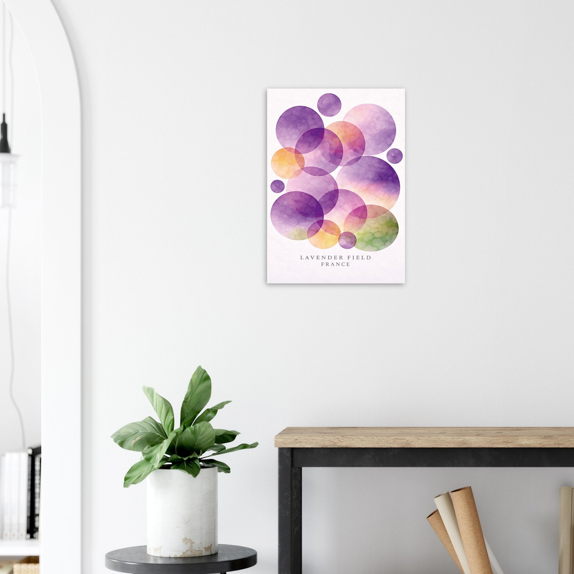 A vertical watercolor art print featuring overlapping purple, pink, orange, and green circles on a white background with the text 'LAVENDER FIELD, FRANCE' at the bottom, displayed on a white wall above a wooden table and next to a potted plant.