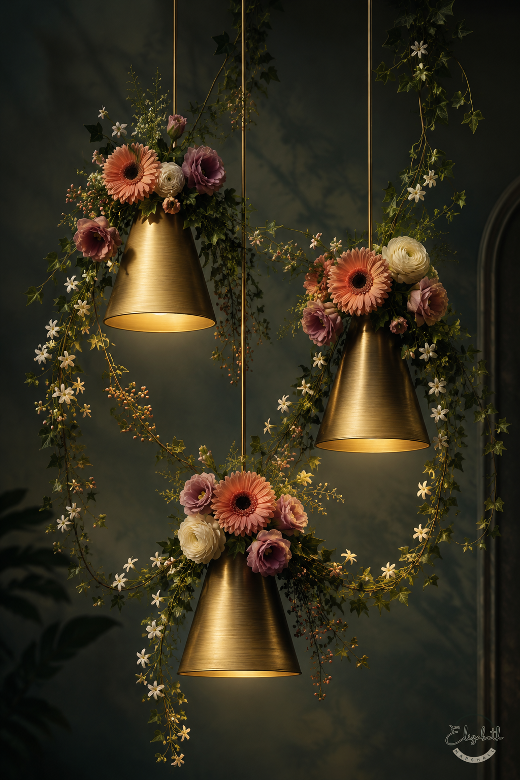 Three hanging brass pendant lights with warm glowing bulbs, decorated with floral arrangements of orange, pink, and white flowers with green vines, set against a dark textured wall.