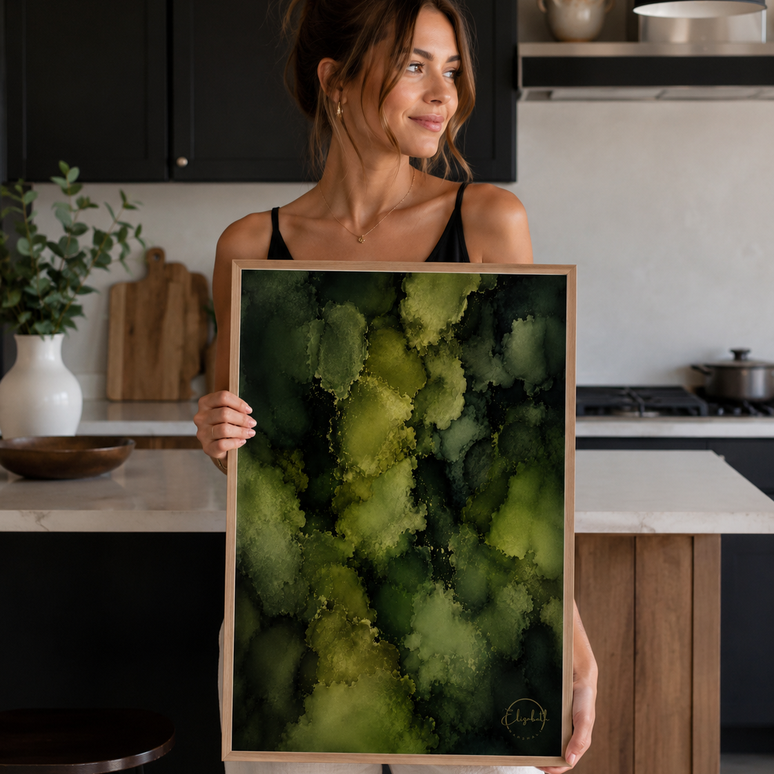 A woman in a kitchen setting holds up a framed abstract art print featuring textured splashes of olive green, lime, and dark forest green.