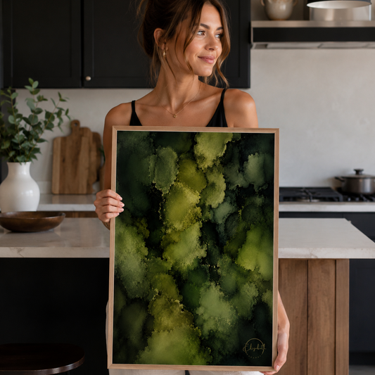 A woman in a kitchen setting holds up a framed abstract art print featuring textured splashes of olive green, lime, and dark forest green.