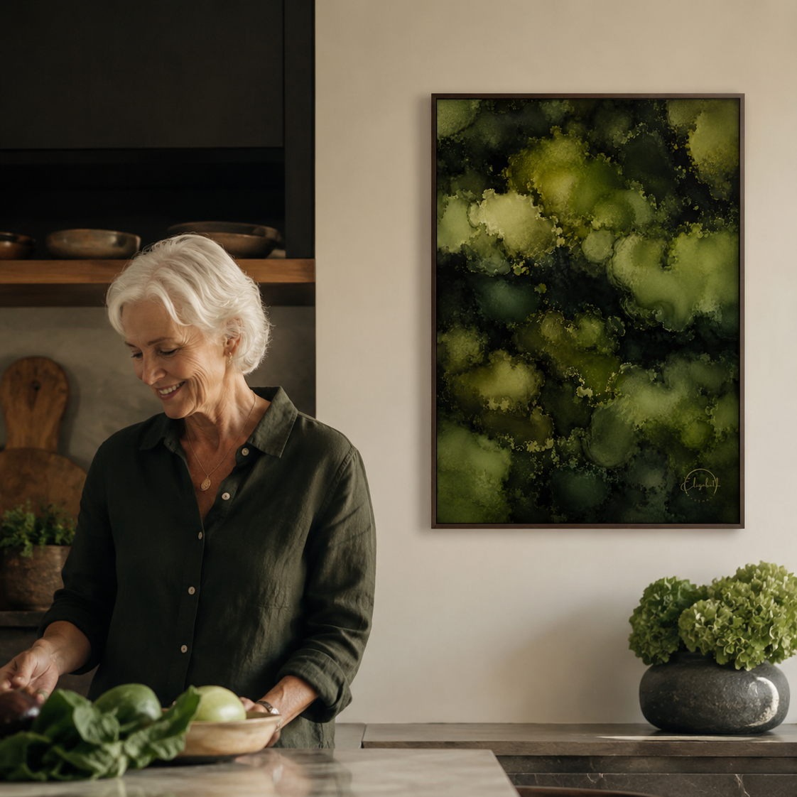 A woman with short white hair stands at a kitchen island, smiling while preparing vegetables. On the wall above the counter hangs a framed abstract artwork in shades of green and black.
