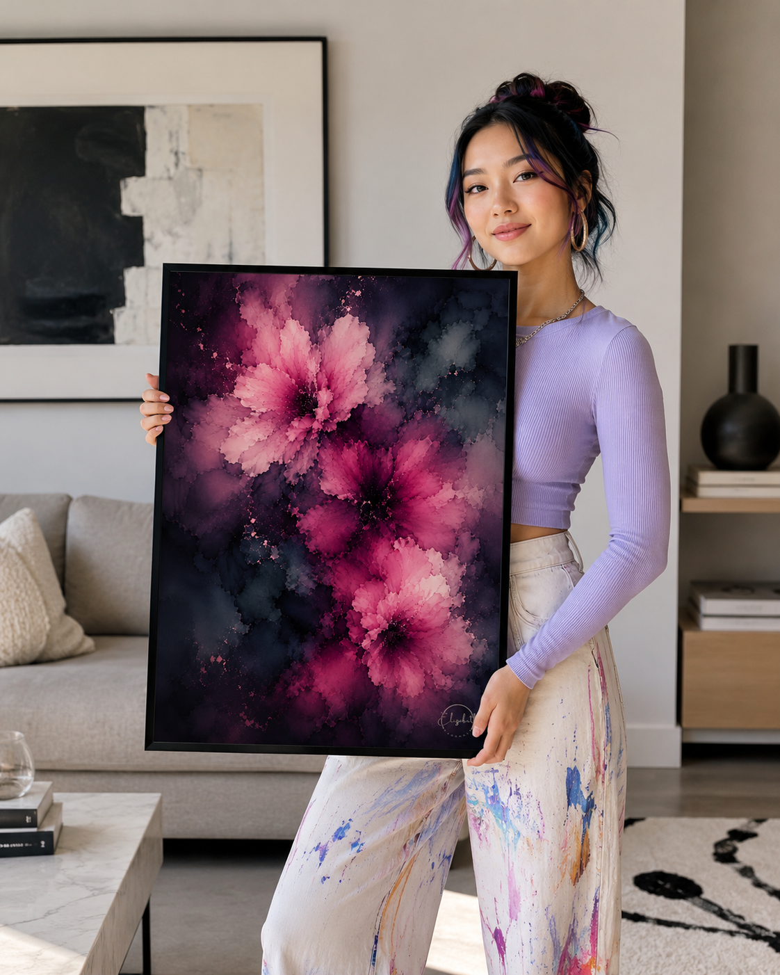 A woman in a lavender long-sleeve top and paint-splattered pants holds up a large framed abstract floral painting featuring vibrant pink and magenta flowers against a dark purple and black background, set in a modern living room environment.