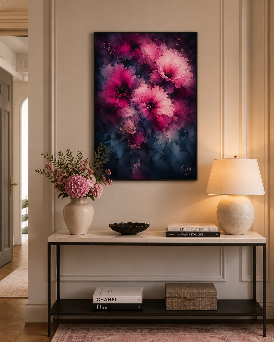 A vertical abstract floral canvas print with vibrant pink and purple flowers on a dark navy background, styled on a beige wall above a console table with a lamp, flowers, and books.