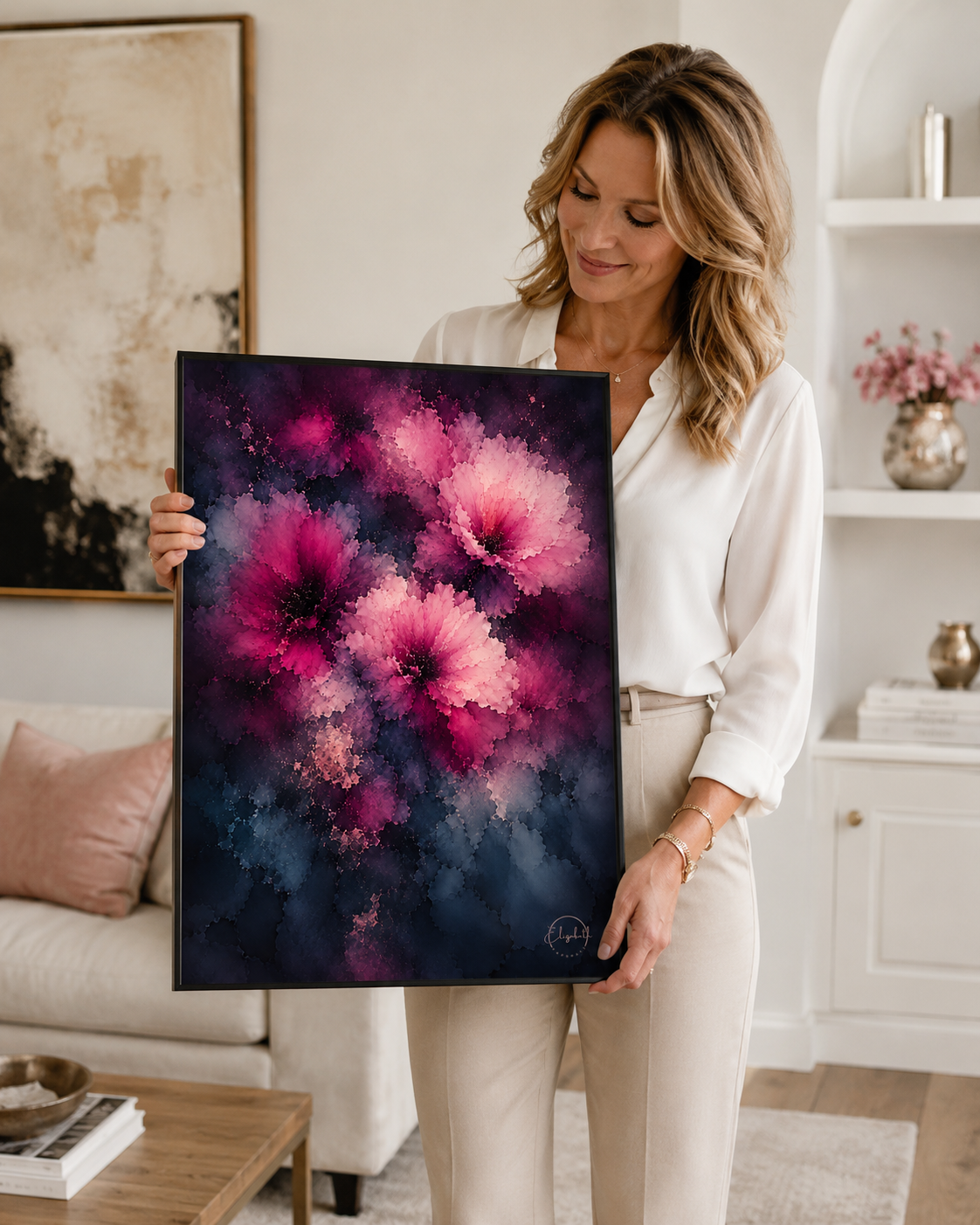 A woman in a white blouse and beige pants holds up a framed abstract floral painting featuring vibrant pink and magenta flowers against a dark navy and purple background, set in a bright, modern living room.