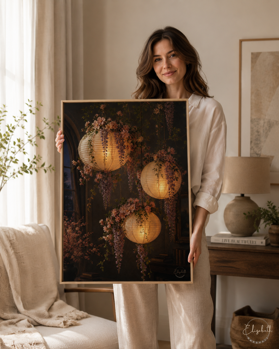 A woman in a cream-colored outfit holds up a large framed art print depicting three glowing paper lanterns surrounded by wisteria flowers against a dark background, set in a cozy, modern living room.