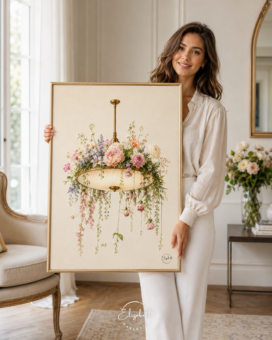A woman in a white blouse and white pants holds up a large framed floral art print featuring a hanging chandelier surrounded by pink, purple, and green flowers. The print has a cream background and a thin gold frame, set against a bright, elegant room with a chair, side table, and mirror.