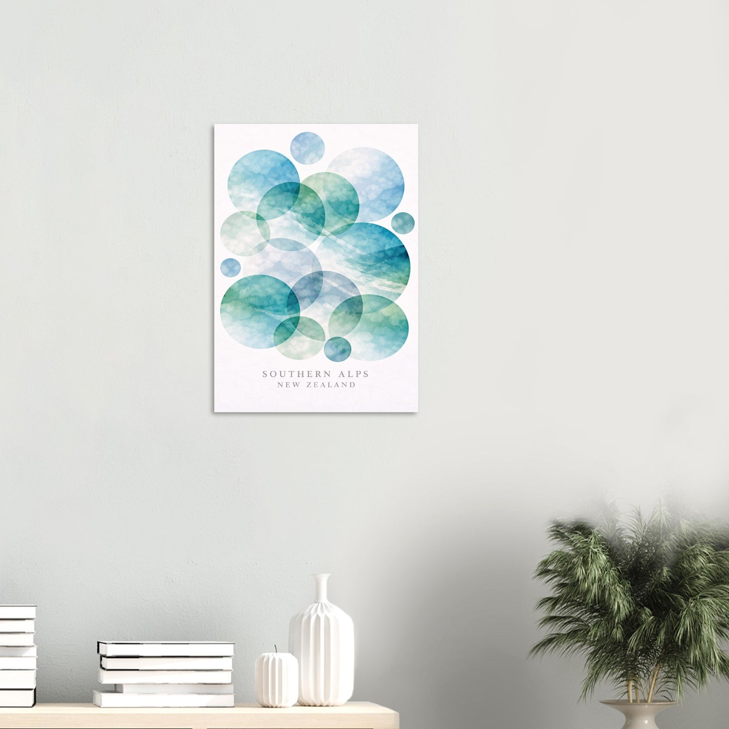 A vertical wall art print featuring overlapping watercolor circles in shades of blue and teal on a white background, with the text 'SOUTHERN ALPS NEW ZEALAND' near the bottom. The print is displayed on a light gray wall above a shelf with books, a white vase, and a potted plant.