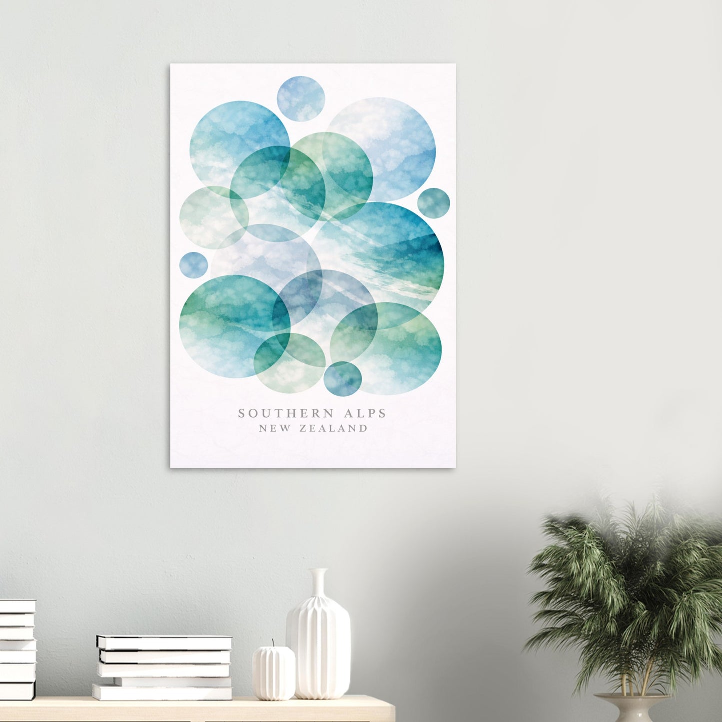 A vertical wall art print featuring overlapping watercolor circles in shades of blue, teal, and green on a white background, with the text 'SOUTHERN ALPS NEW ZEALAND' near the bottom. The print is displayed on a light gray wall above a shelf with books, a white vase, and a potted plant.
