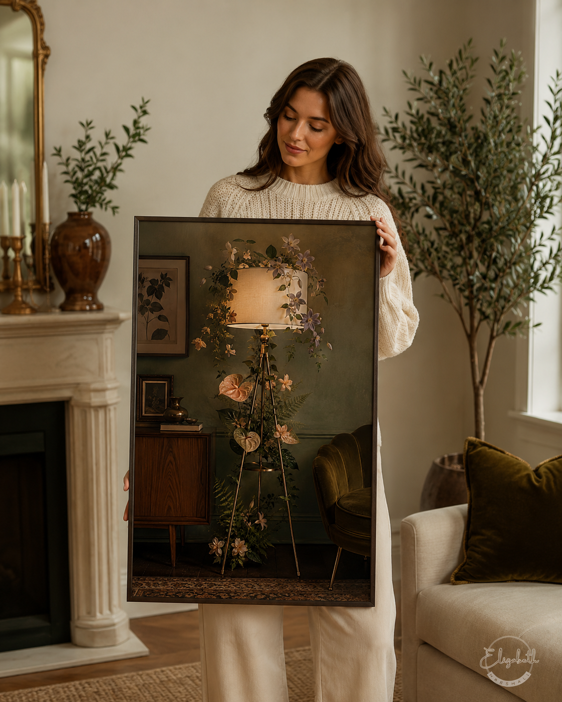 A woman in a cream knit sweater holds up a large framed art print depicting a cozy room scene with a floral floor lamp, set in a beautifully decorated living room with a fireplace and potted plants.