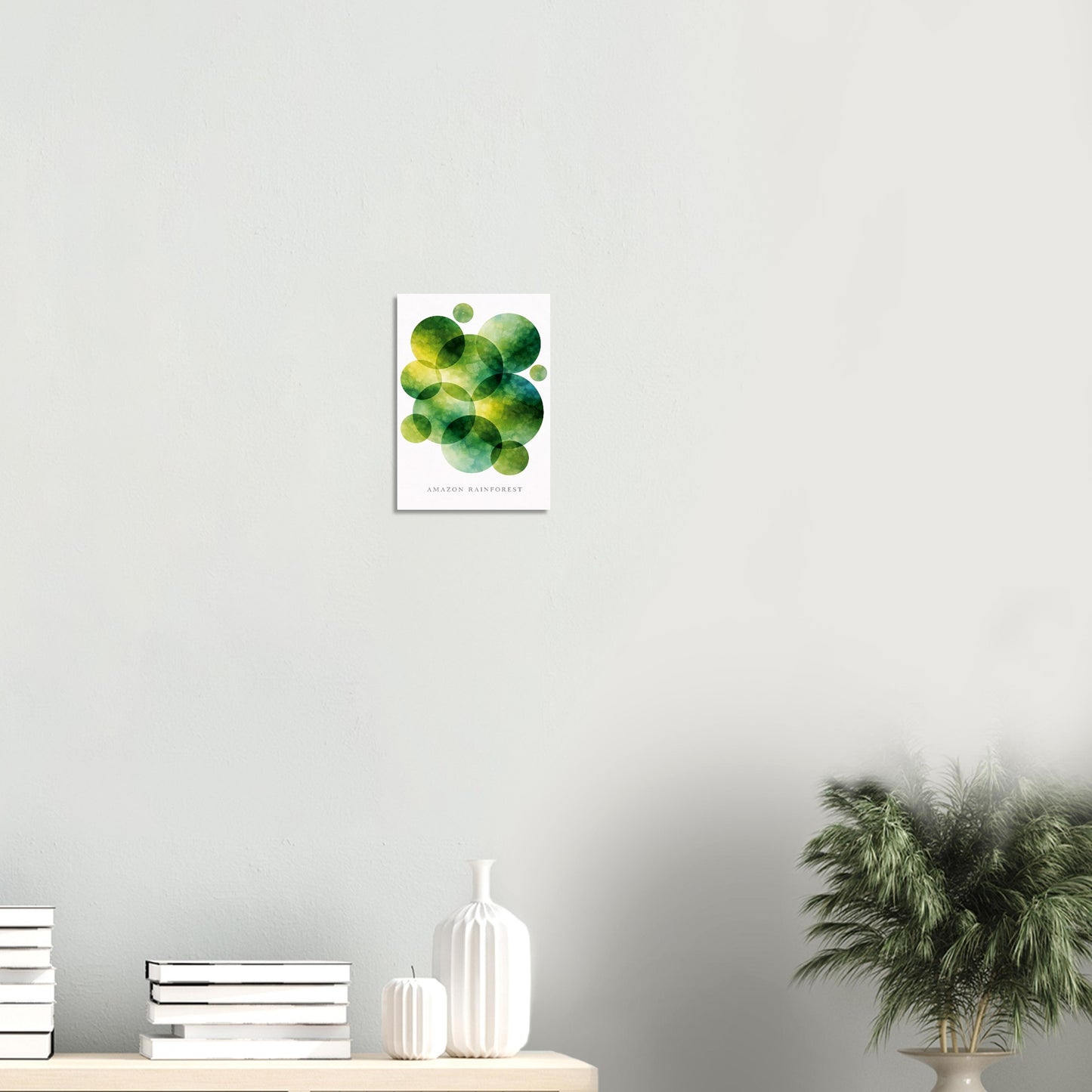 A vertical art print featuring green and yellow watercolor circles on a white background, displayed on a light gray wall above a wooden shelf with books, a white vase, and a potted plant.
