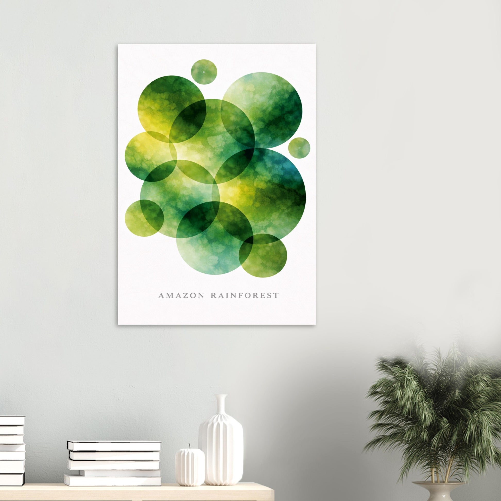 A vertical wall art print featuring overlapping green and yellow watercolor circles with the text 'AMAZON RAINFOREST' below, displayed on a light gray wall above a console table with books, a vase, and a potted plant.