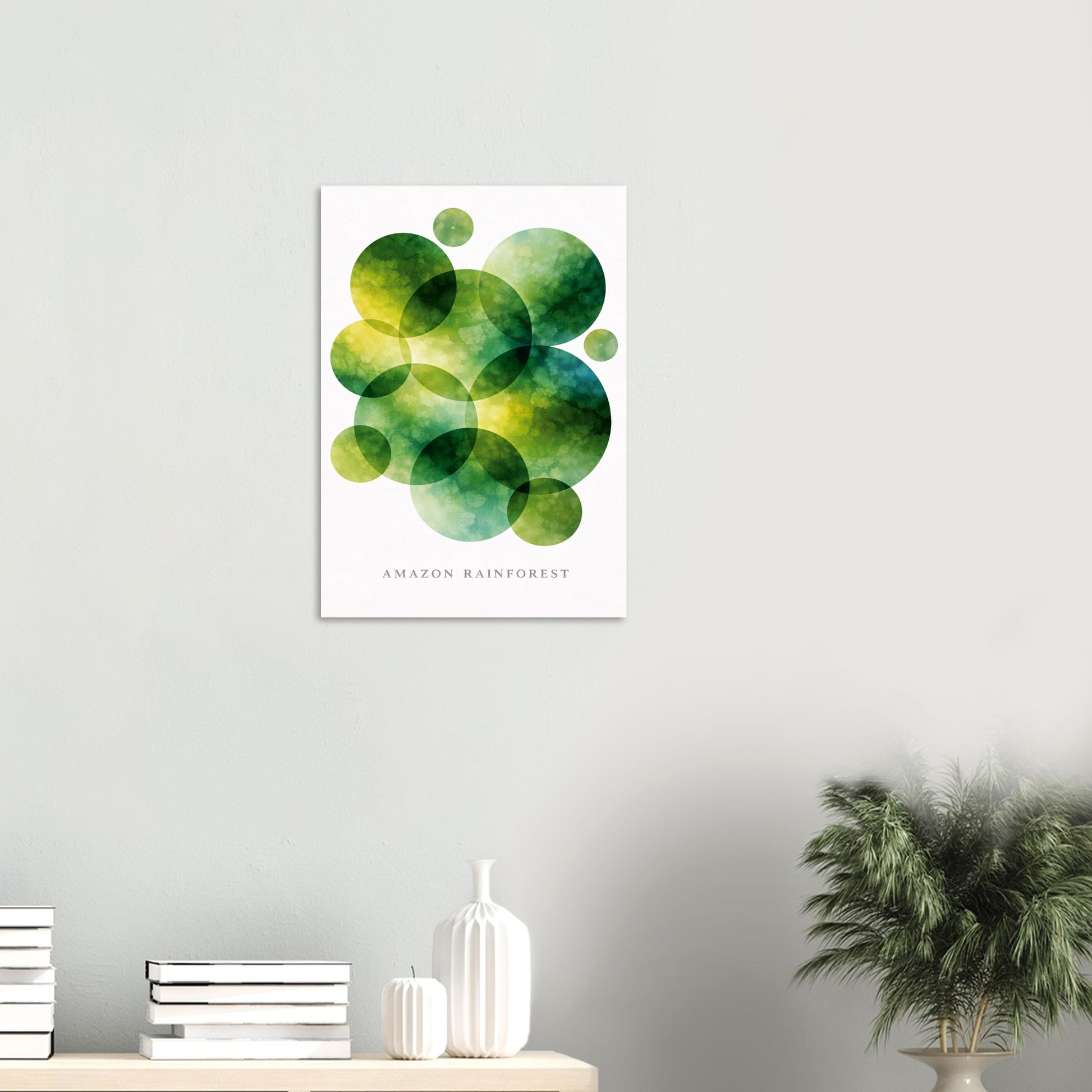 A vertical wall art print featuring overlapping green and yellow watercolor circles on a white background, with the text 'AMAZON RAINFOREST' near the bottom. The print is displayed on a light gray wall above a shelf with books, white vases, and a potted plant.