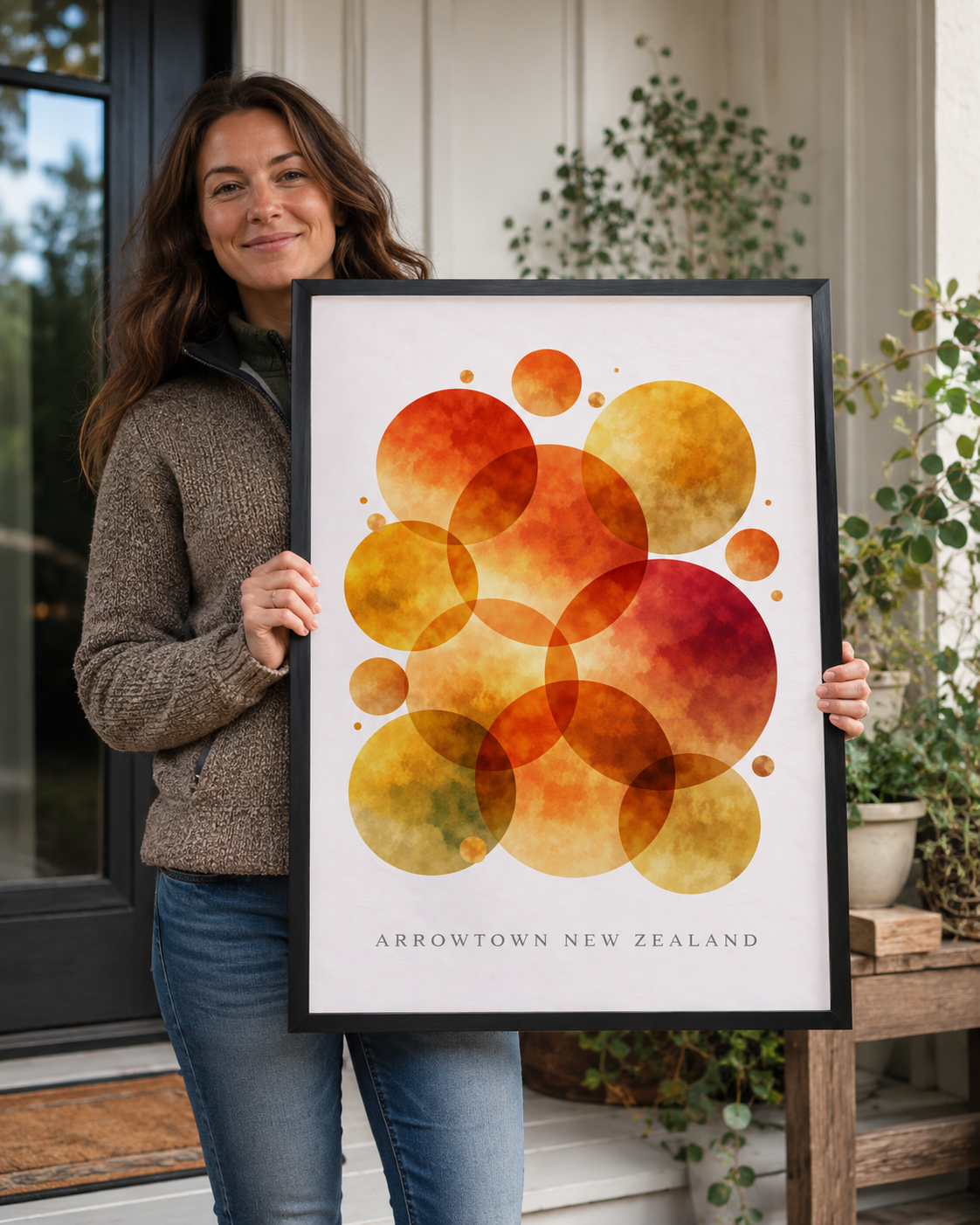 A woman holds a framed art print featuring overlapping watercolor circles in orange, yellow, and red on a white background, with the text 'ARROWTOWN NEW ZEALAND' at the bottom. She is standing in front of a house exterior with plants.