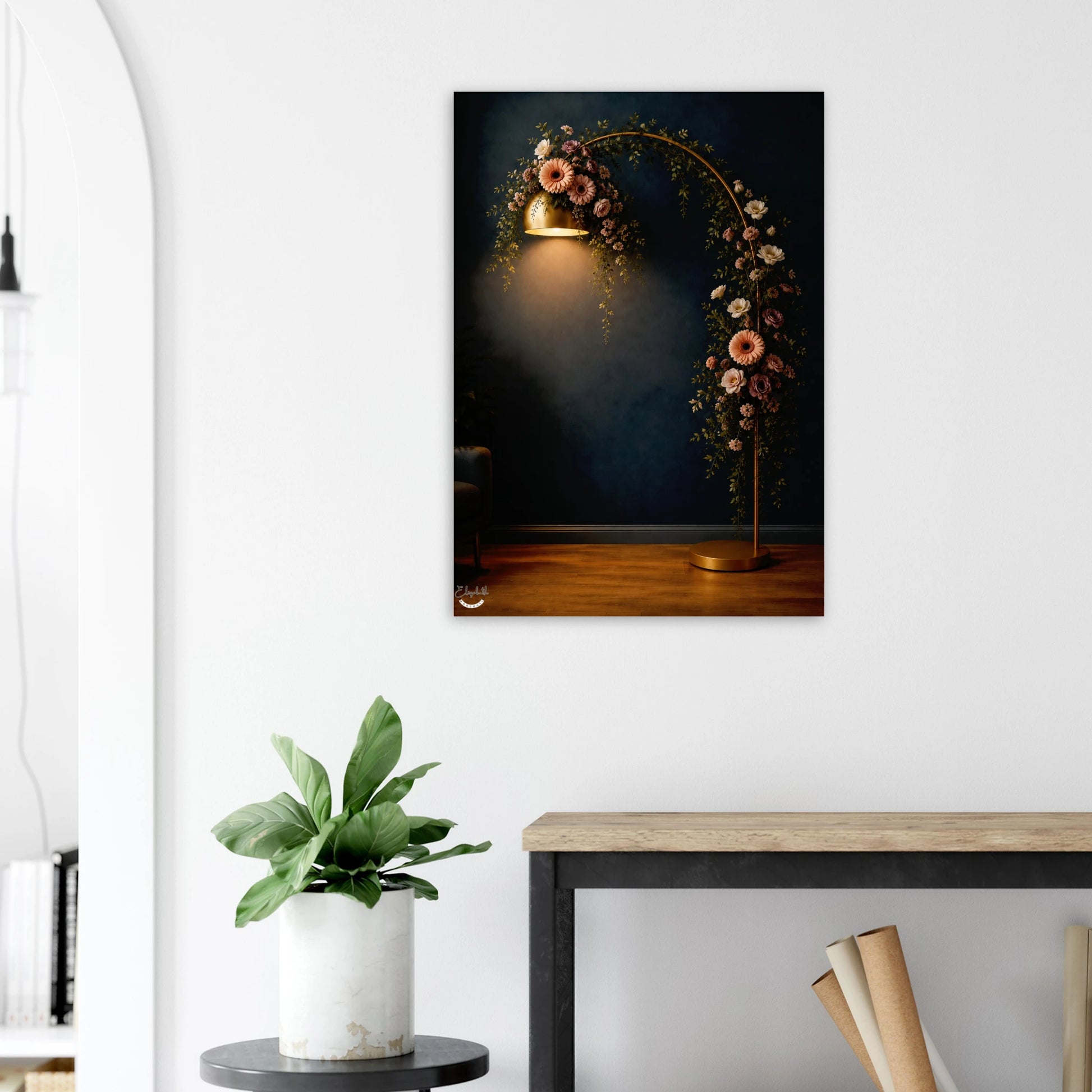 A vertical wall art print depicting a glowing brass floor lamp draped with pink and white flowers against a dark blue background, shown hanging on a white wall above a console table with a potted plant.