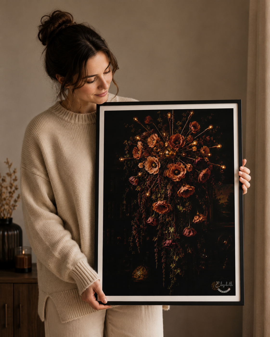 A woman in a beige knit sweater holds a large framed art print featuring a dark, moody floral composition with glowing warm highlights. The print has a black background, a white mat, and a thin black frame, shown in a warm, neutral-toned indoor setting.
