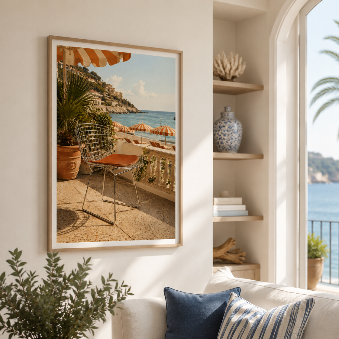 A framed art print depicting a sunny coastal balcony scene with a chair, plants, and the sea, styled on a cream wall in a bright living room above a sofa with blue and striped pillows.