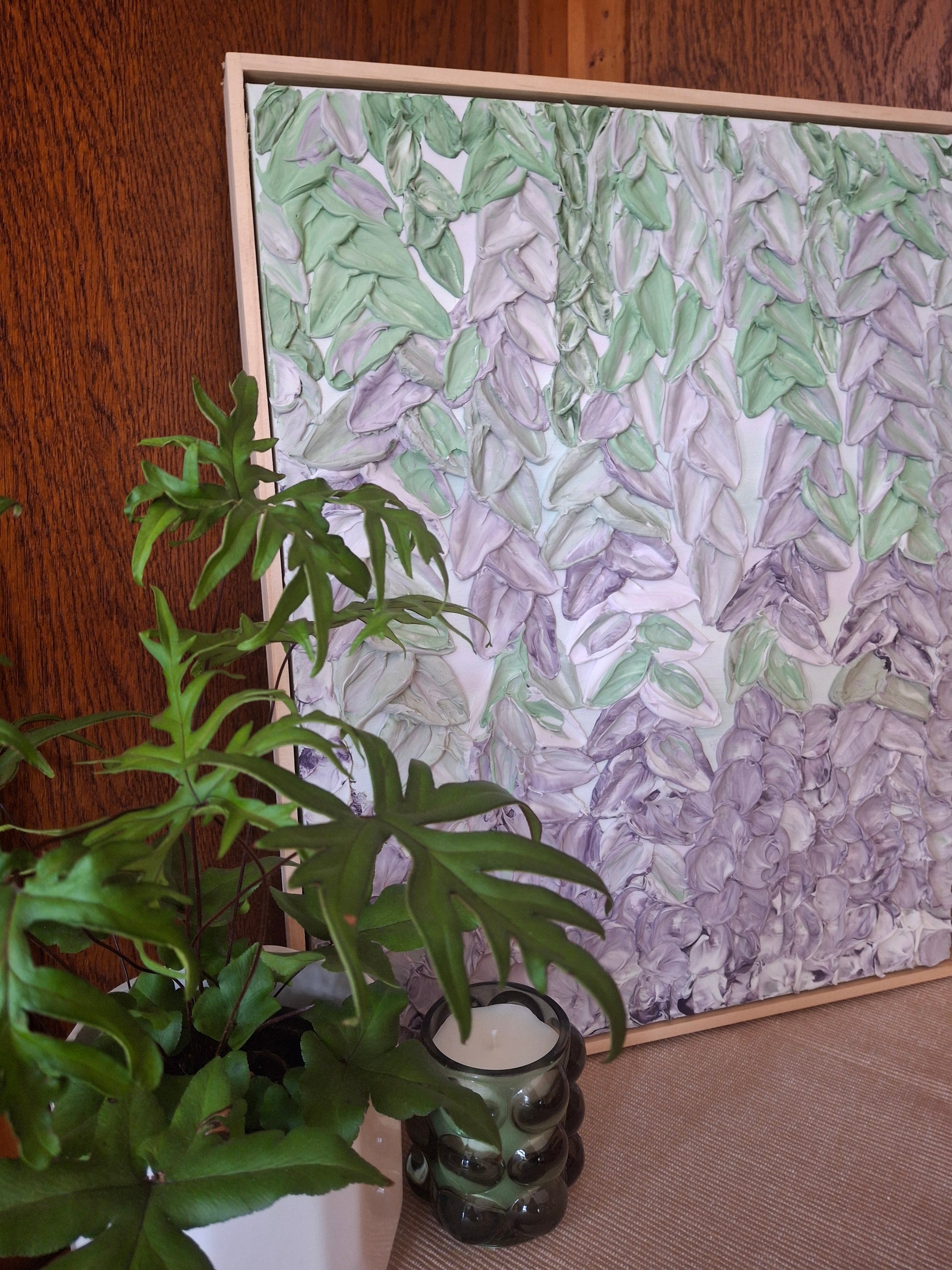 A framed artwork featuring a textured, layered pattern of green and lavender-gray leaves on a white background, styled on a wooden table next to a green plant and a glass candle holder.