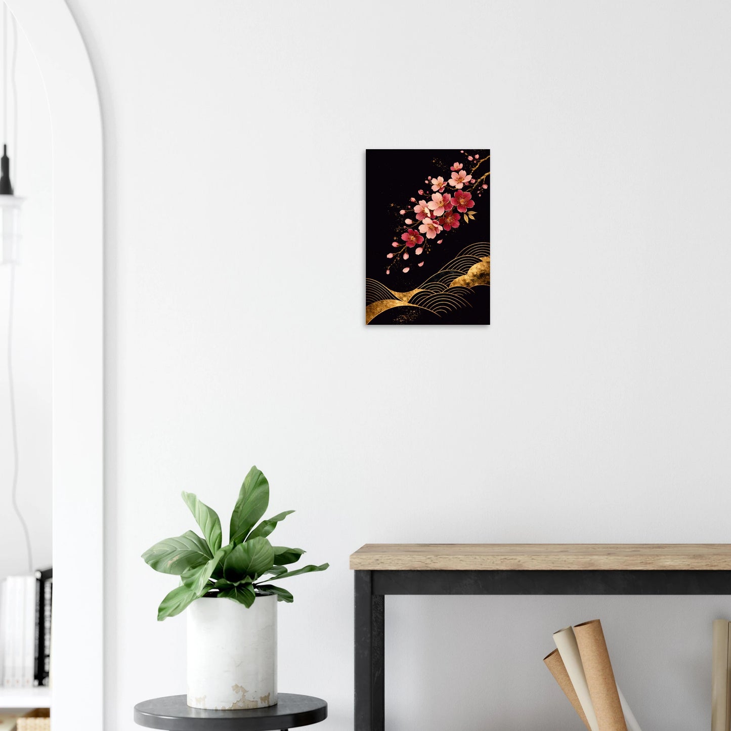 A vertical wall art print featuring pink cherry blossoms and gold abstract waves on a black background, displayed on a white wall above a console table with a potted plant.