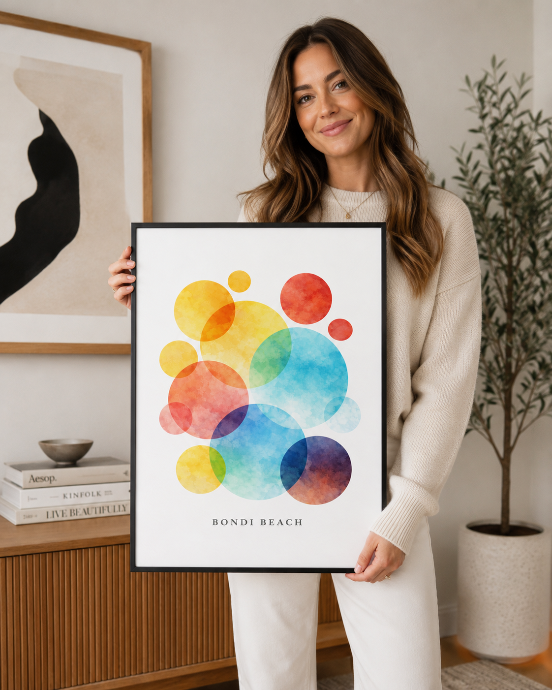A woman in a cream knit sweater and white pants holds a framed art print featuring overlapping watercolor circles in yellow, orange, red, blue, and purple, with the text 'BONDI BEACH' at the bottom. The setting is a bright, modern room with a wooden console table, a potted plant, and another framed print on the wall.