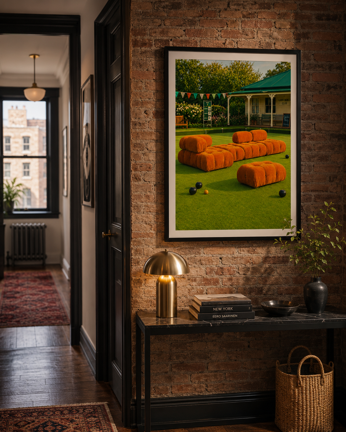 A framed art print depicting a vibrant green lawn with bright orange modular seating and a porch in the background, displayed on a brick wall above a console table in a modern interior hallway.