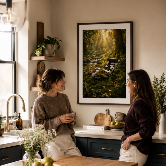 A large framed art print depicting a lush green forest stream is displayed on a beige wall in a modern kitchen, with two women standing nearby and engaging in conversation. The Chaise Went Wild and honestly, it was only a matter of time. A mid-century chaise lounge, dark frame and black and white upholstery, sits deep in an enchanted forest, moss creeping up its legs, tiny white wildflowers pressing in from every side, golden light filtering down through the canopy above. It is not lost. It is exactly where