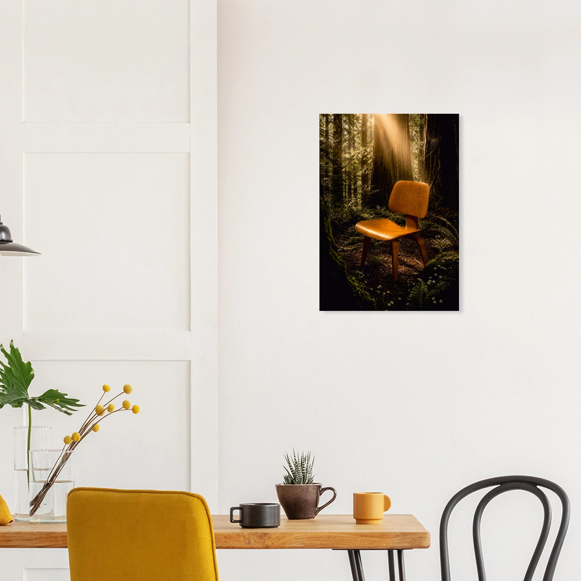 A vertical canvas wall art print depicting a mid-century modern yellow chair in a dark, sunlit forest, displayed on a white wall above a dining table in a modern room setting.