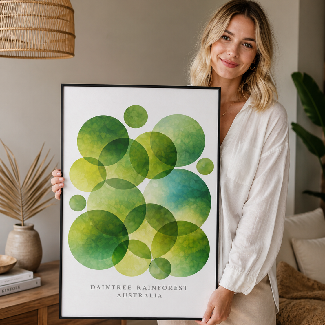 A woman in a white shirt and beige pants holds up a framed art print featuring overlapping green and blue watercolor circles with the text 'DAINTREE RAINFOREST AUSTRALIA' at the bottom.