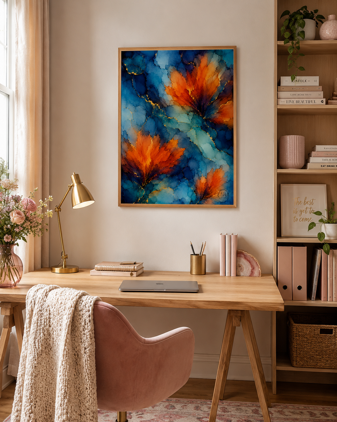 A framed abstract wall art print featuring vibrant orange and red floral-like forms over a deep blue background, displayed in a warm, modern home office setting above a wooden desk with a pink chair.