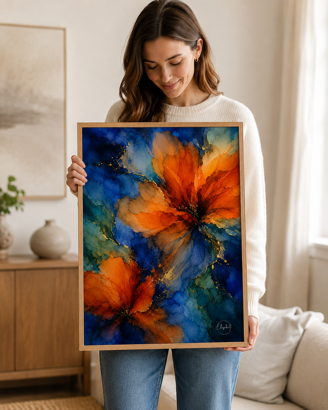 A woman in a cozy living room setting holds up a framed abstract floral painting. The artwork features vibrant orange flowers set against a deep blue and teal background, enclosed in a light natural wood frame.