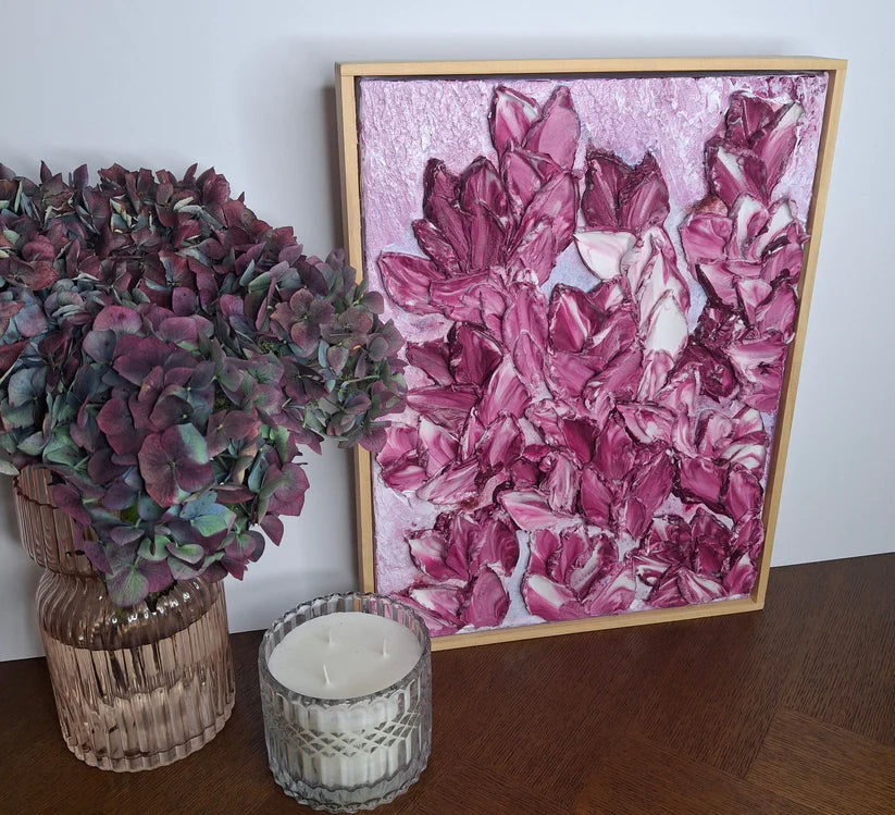 A framed abstract painting featuring textured pink and purple brushstrokes, styled on a wooden table next to a glass vase with dried purple flowers and a lit white candle.