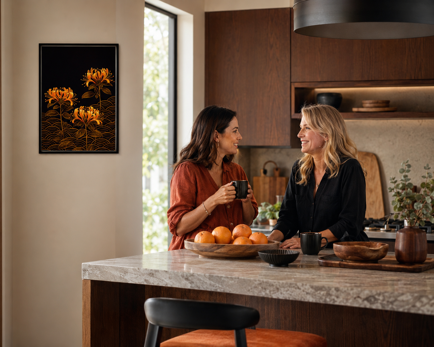A framed wall art print featuring orange flowers on a black background hangs on a beige wall in a modern kitchen. Two women are standing at a marble kitchen island, holding mugs and smiling, with oranges and bowls on the counter.