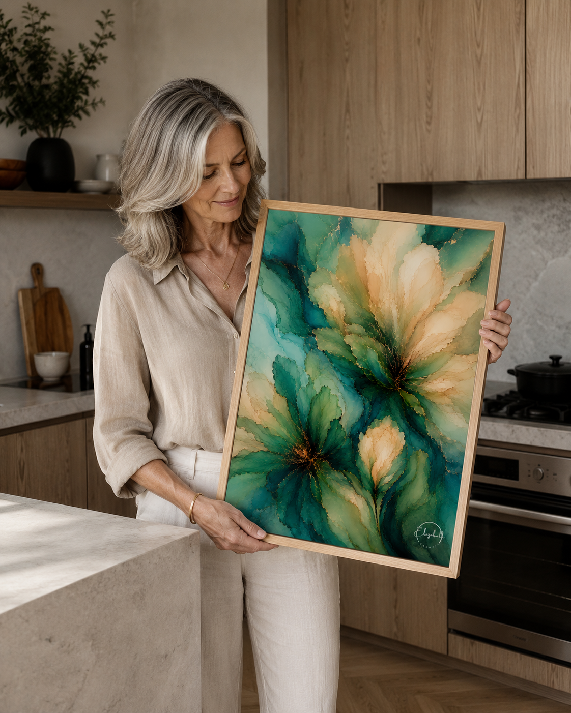 A woman with gray hair stands in a modern kitchen holding a framed abstract floral painting with teal, green, and cream tones.